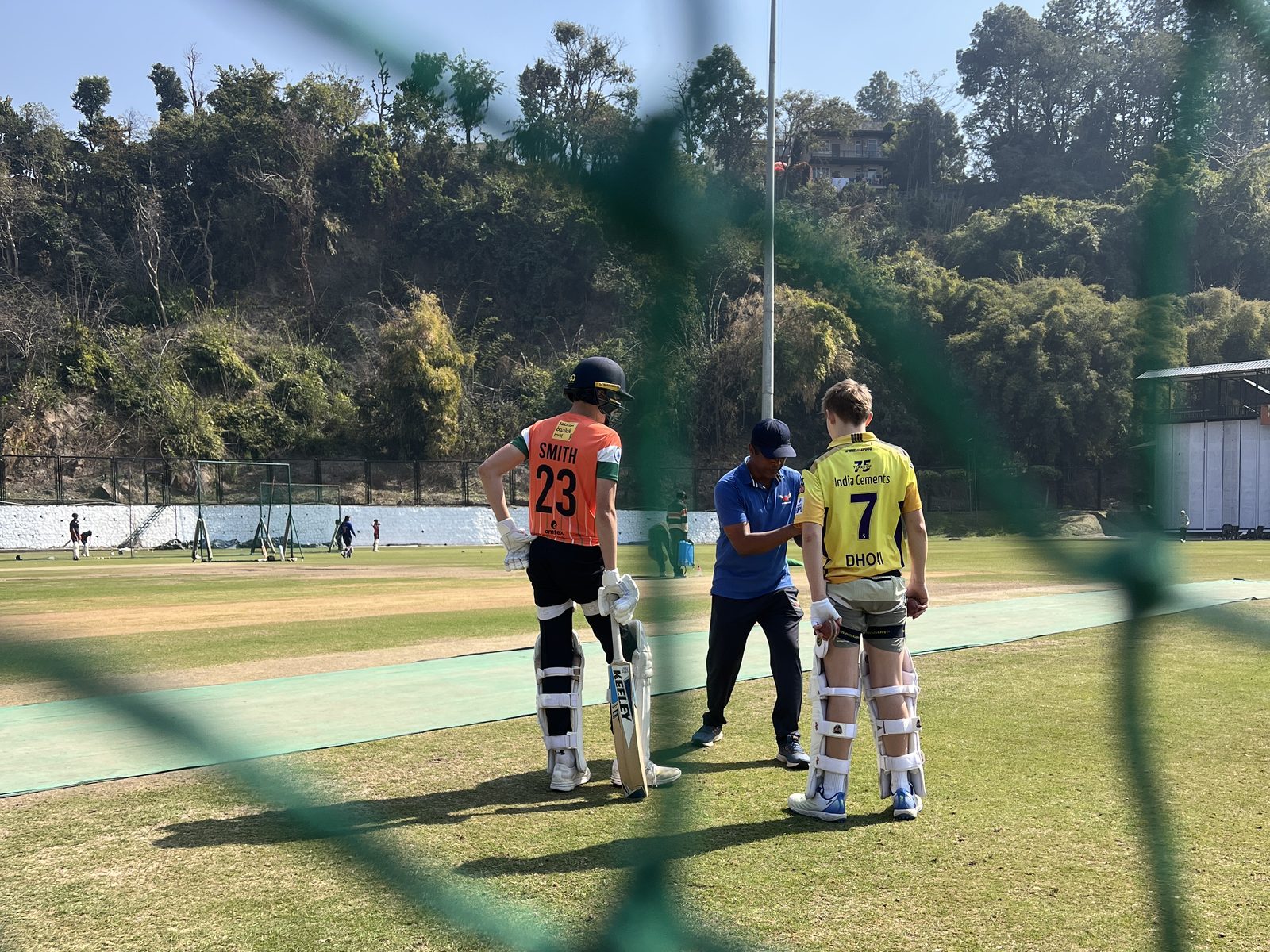 A coach working with two batters, seen through the nets